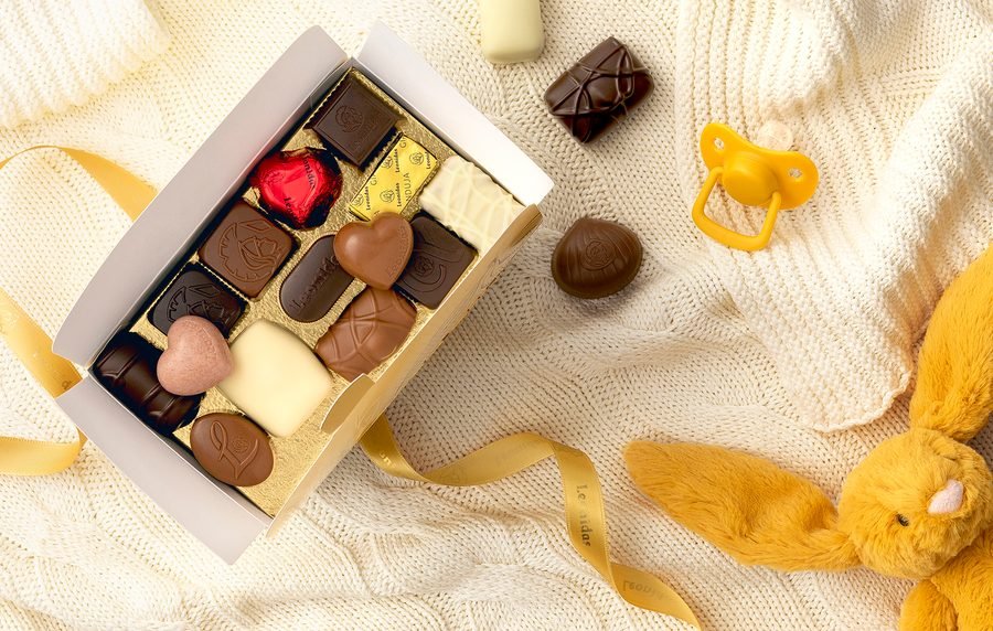 A box of Leonidas pralines on a baby blanket, with a soft toy and a pacifier